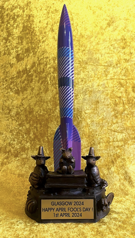 An ornamental trophy celebrating April Fool's Day in Glasgow for the year 2024. The trophy features a large, vertically standing, patterned rocket in hues of purple and blue with tartan pattern. The base is a dark, metallic color, depicting three armadillos, two in traffic cone hats flank a centered armadillo sitting atop a book. Center, a plaque reads 'GLASGOW 2024 HAPPY APRIL FOOLS' DAY! 1st APRIL 2024' against a textured gold background.